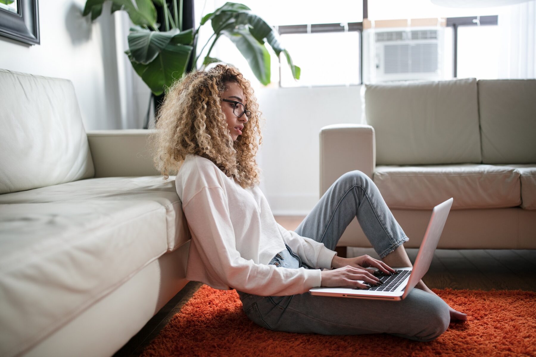 lady with glasses sitting on the floor with a computer leaning on a grey couch